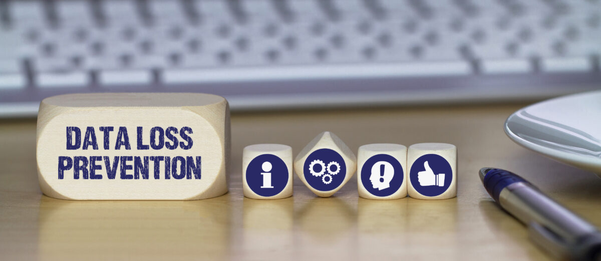 Wooden block with "Data Loss Prevention" written on it, accompanied by smaller blocks featuring icons for information, gears, a person with an exclamation mark, and a thumbs-up, placed on a desk with a pen and keyboard in the background.