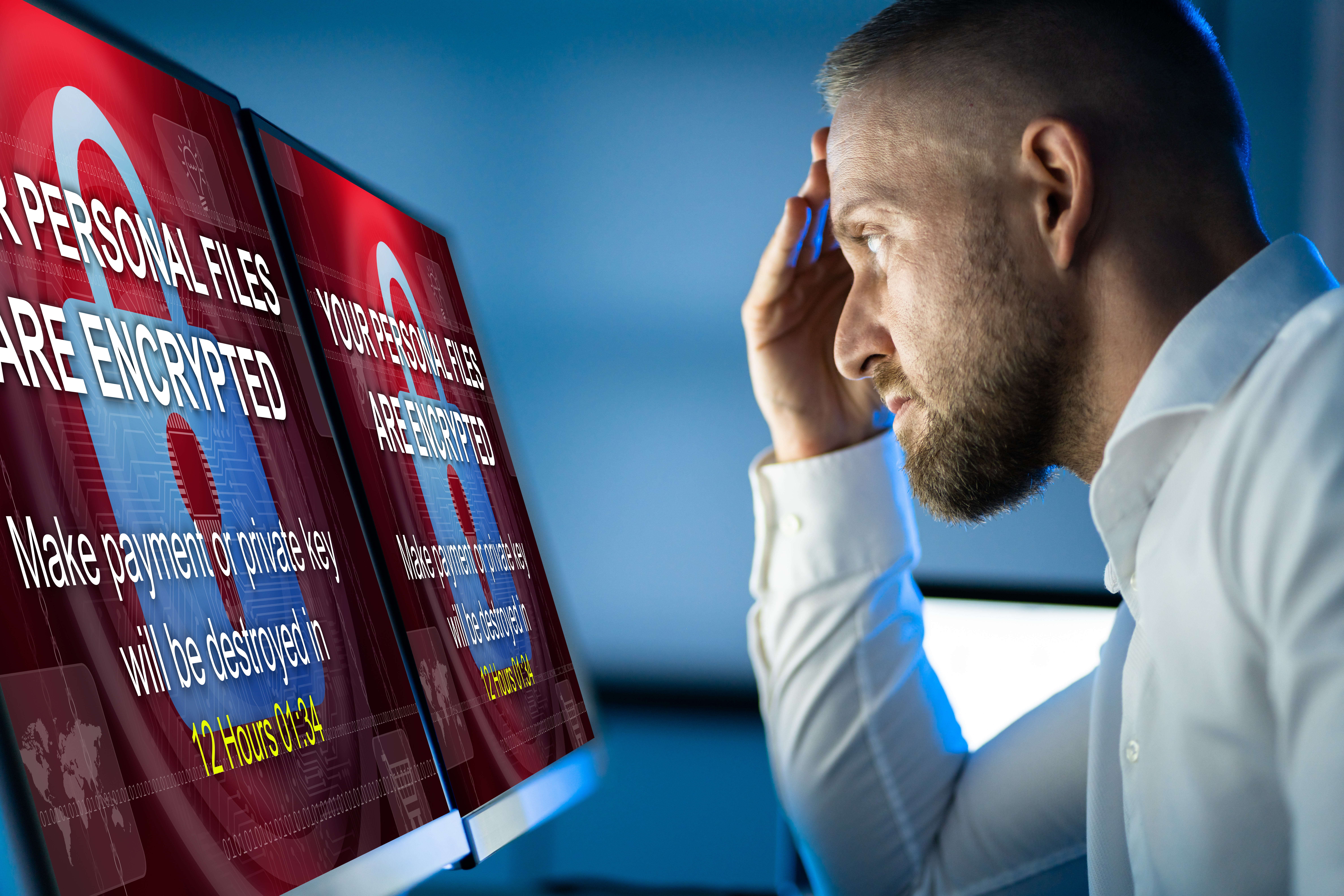 Man in a white shirt sitting at a desk, looking stressed while viewing dual monitors displaying a ransomware message stating 'Your personal files are encrypted. Make payment or private key will be destroyed in 12 hours